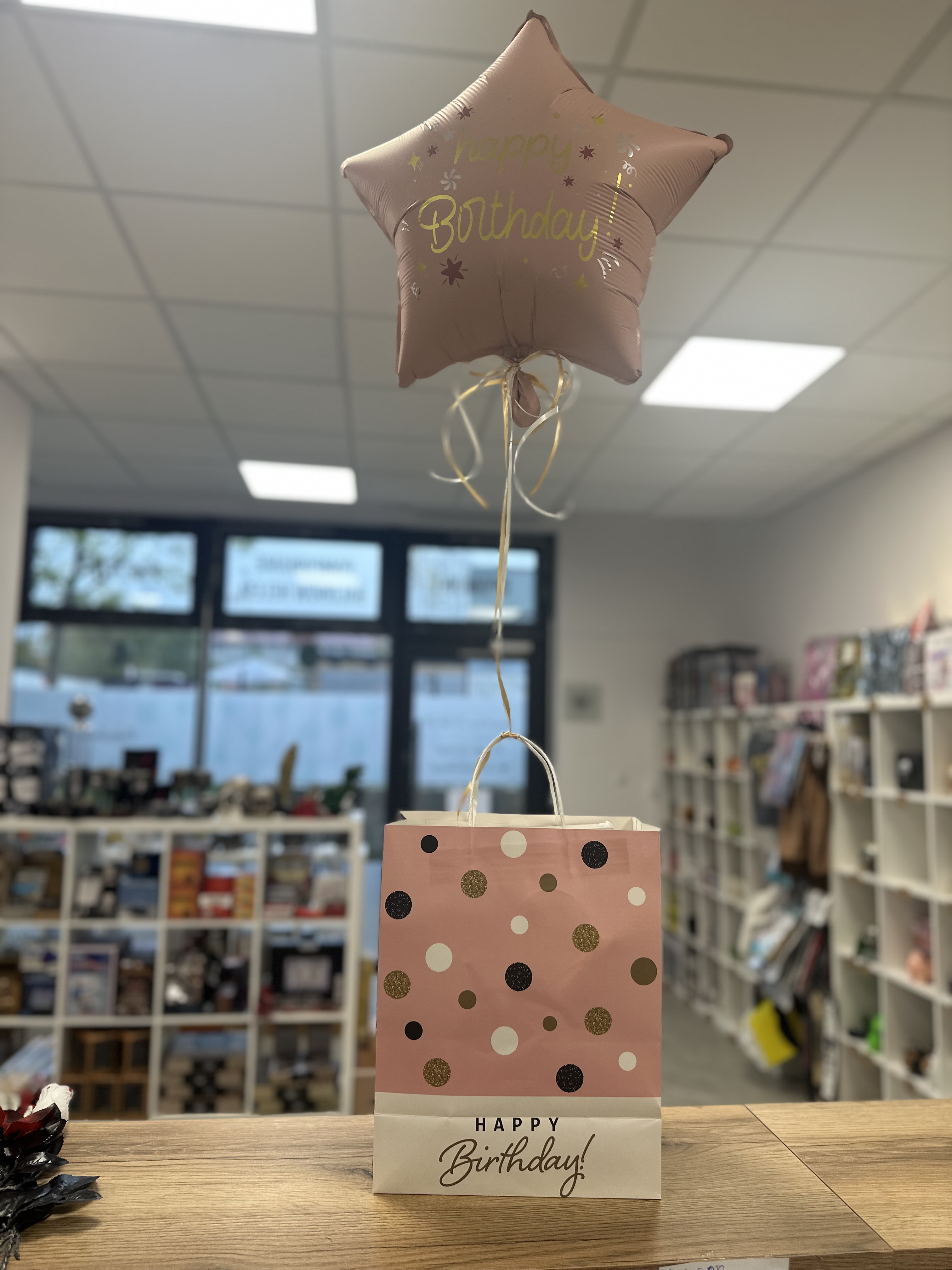 Prezent zapakowany w ozdobną torbę prezentową, do której przyczepiony jest balon z helem w kształcie gwiazdki z napisem Happy Birtday. Torebka i gwiazdka w kolorze różowym.